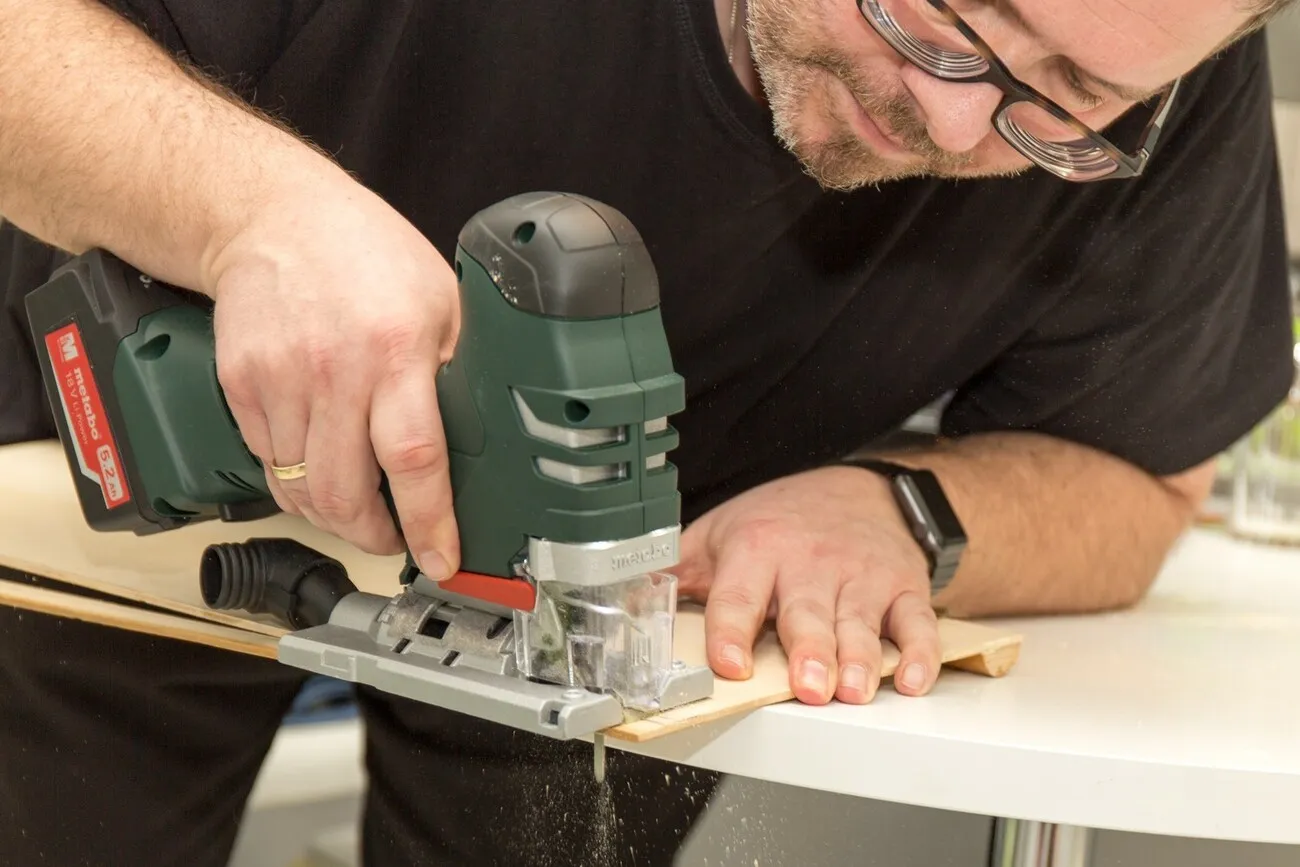 Ein Mann sägt mit der Metabo Stichsäge am Tischende eine Multiplexplatte.